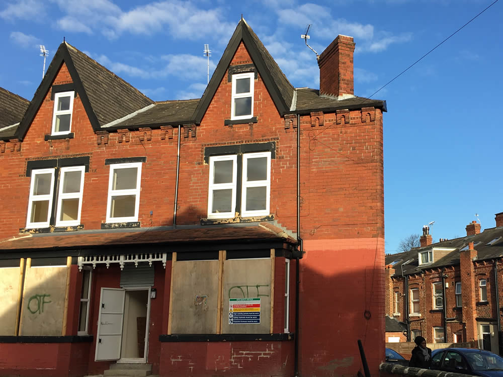A large brick house with pointed roof and large boarded up windows downstairs